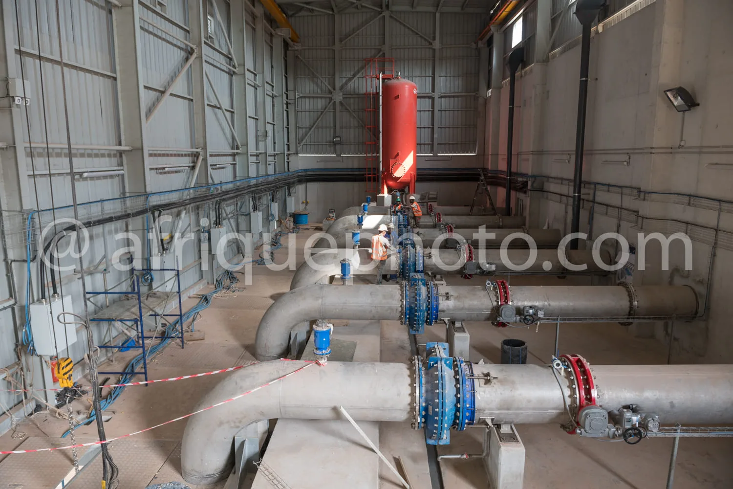 Africa photo — Vue d’un atelier industriel montrant une complexe tuyauterie en métal inoxydable, des vannes bleues et rouges, ainsi qu’un grand réserv