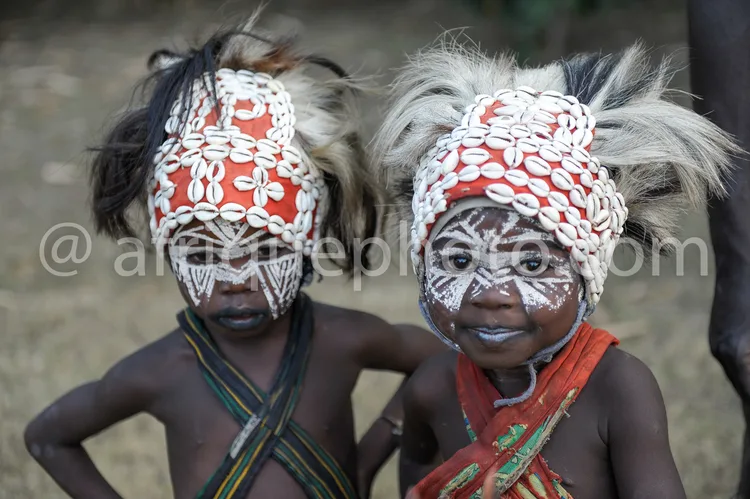 Africa photo — Deux enfants souriants vêtus de parures traditionnelles composées de coquillages blancs et d’un bandeau rouge, avec des motifs peints e