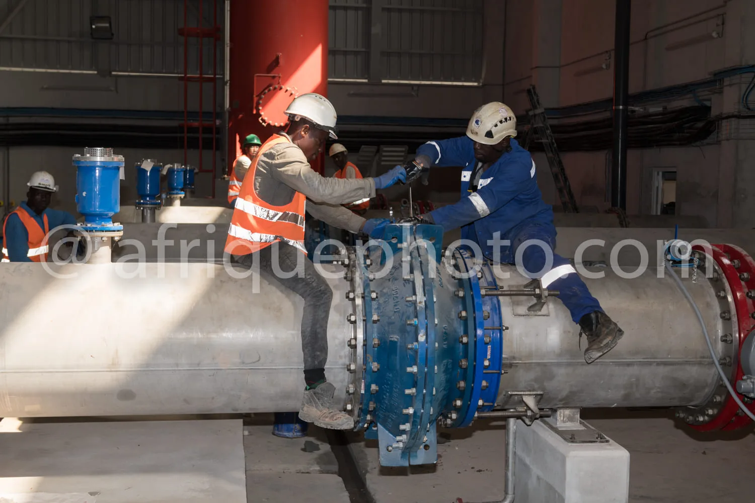 Africa photo — Deux techniciens en combinaison de travail, casques de protection et gilets de sécurité orange ou bleu, serrent manuellement une bride