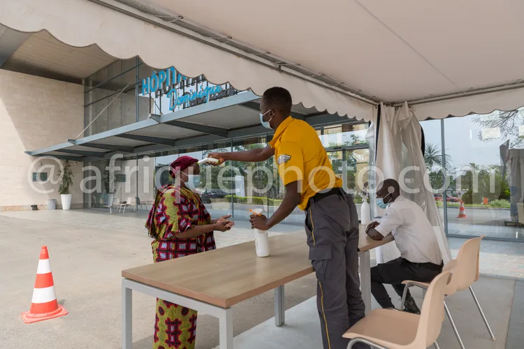 Africa photo — Un homme en tenue jaune et masque distribue un gobelet en plastique à une femme assise à une table en extérieur, sous une tente blanche