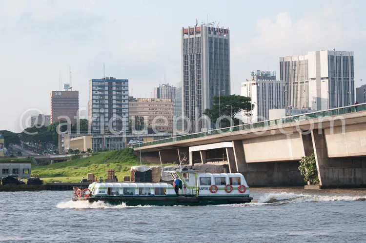 Africa photo — Un bac fluvial blanc avec des passagers et des véhicules à bord traverse un fleuve calme. À l’arrière-plan, une ville moderne avec des