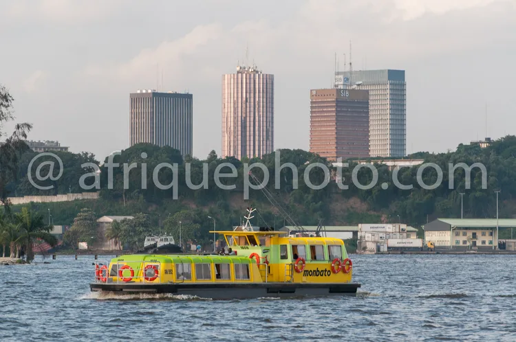 Africa photo — Un bateau jaune vif nommé Monbato navigue sur une étendue d’eau calme, avec en arrière-plan une skyline urbaine composée de plusieurs i