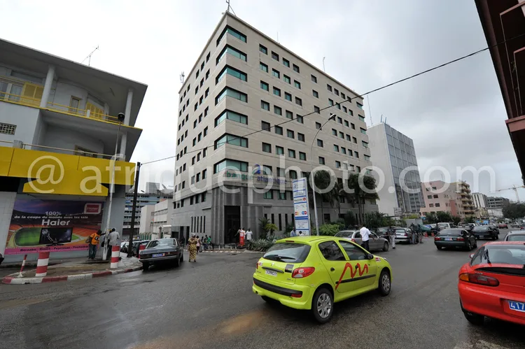 Africa photo — Un immeuble de quatre étages en béton clair avec des balcons et des fenêtres alignées, situé au carrefour d’une rue animée. À gauche, u