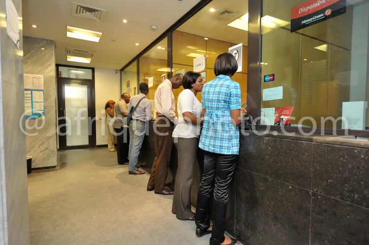 Africa photo — Six personnes debout en file devant un guichet bancaire vitré, avec un panneau indiquant 'Disponible' et des services comme le mobile b