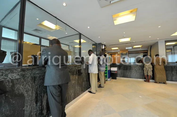 Africa photo — Quatre personnes se tiennent près d’un comptoir de guichet bancaire moderne en marbre gris, éclairé par des néons au plafond. Un homme