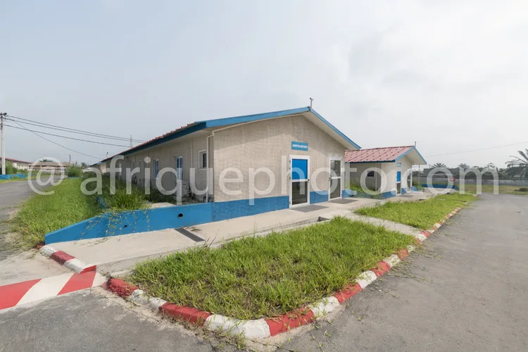 Africa photo — Un petit bâtiment administratif en bordure d’une route, avec une façade beige et des bandes bleues horizontales. Une entrée principale