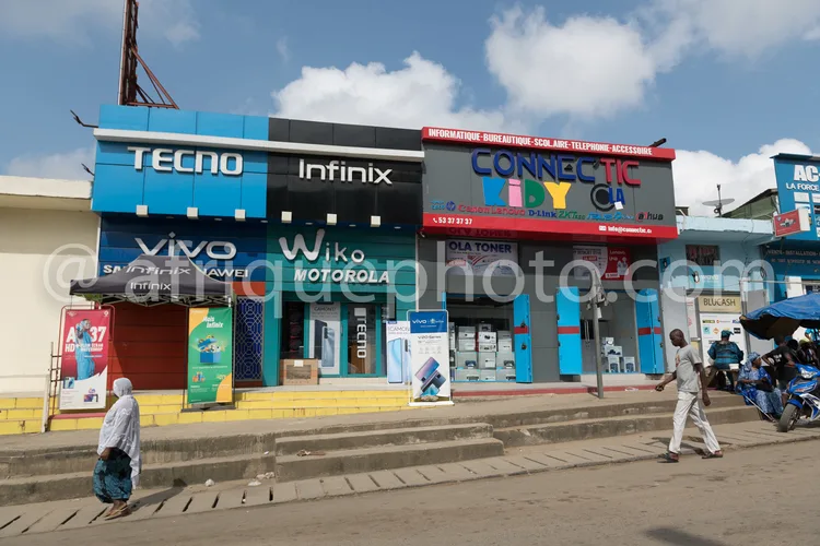 Africa photo — Une rue commerçante avec plusieurs boutiques spécialisées en téléphonie et électronique, affichant des enseignes comme Tecno, Infinix,