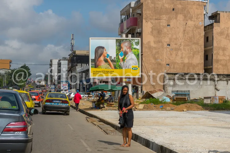 Africa photo — Une femme traverse une rue animée devant un grand panneau publicitaire MTN montrant une femme souriante et un homme en discussion, avec