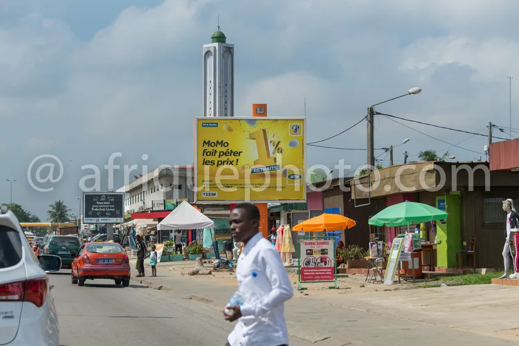 Africa photo — Un homme traverse une rue animée bordée de petits commerces colorés, avec des étals sous parasols, des panneaux publicitaires et une to