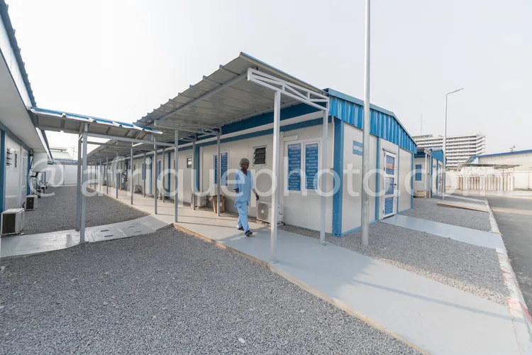 Africa photo — Un professionnel de santé en tenue bleue entre dans un bâtiment médical modulaire composé de conteneurs bleus et blancs, avec une toitu