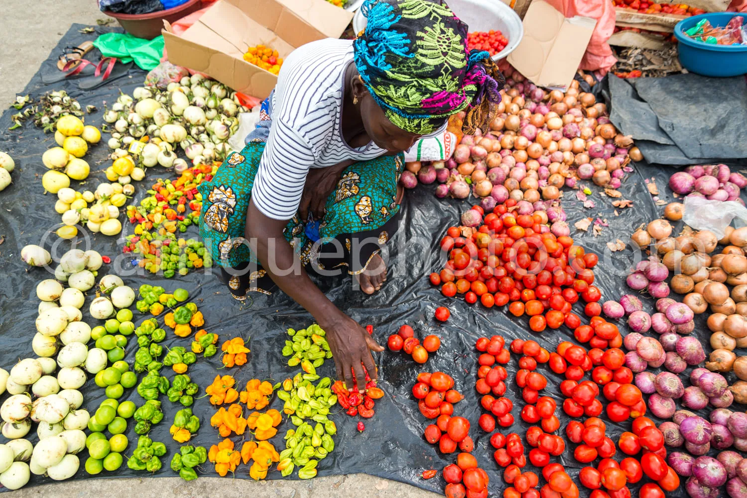 Africa photo — Une femme en tenue traditionnelle, coiffée d’un foulard coloré, est assise sur une bâche noire et trie soigneusement des légumes racine