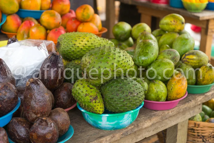 Africa photo — Un étalage coloré de fruits variés : des corossols verts épineux bien en évidence au premier plan, suivis de mangues jaunes et orange,