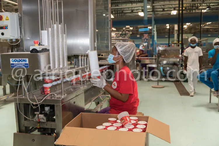 Africa photo — Une employée en tenue de travail rouge et coiffe protectrice manipule une machine industrielle Goldoni pour remplir des gobelets en pla
