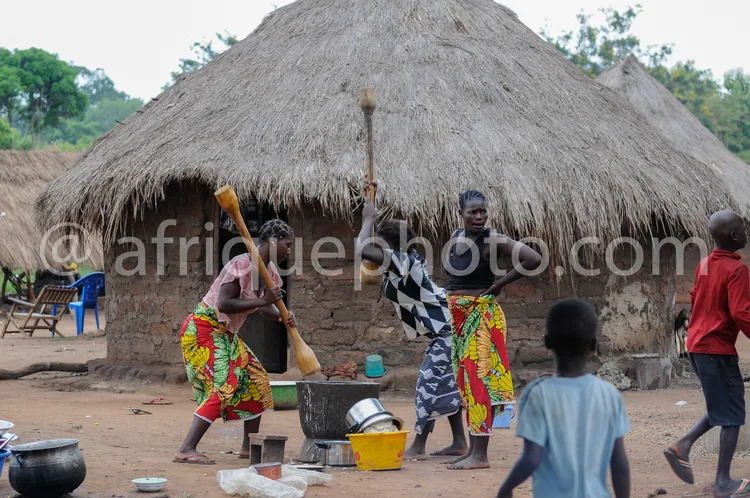 Africa photo — Deux femmes en tenues colorées traditionnelles préparent un repas devant une hutte en chaume, avec des enfants et des ustensiles autour