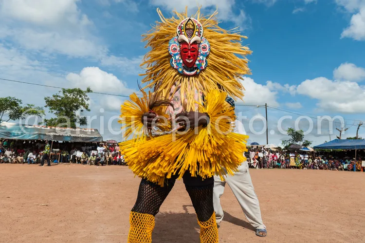 Africa photo — Un danseur porte un imposant masque traditionnel africain en plumes jaunes et motifs rouges et bleus, vêtu d’un costume blanc et noir,