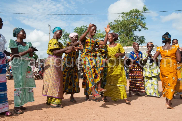 Africa photo — Un groupe de femmes vêtues de robes traditionnelles colorées et de coiffures variées danse en cercle sur un sol en terre battue, sous u