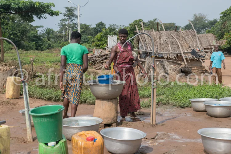 Africa photo — Deux femmes en tenue traditionnelle travaillent ensemble dans un espace extérieur. L’une verse de l’eau dans un grand récipient métalli