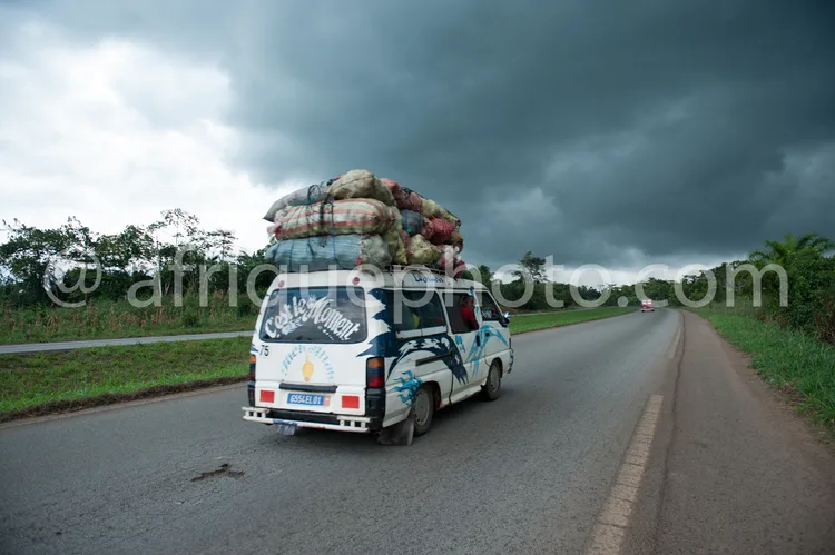 Africa photo — Un véhicule blanc décoré de motifs bleus et argentés roule sur une route goudronnée sous un ciel chargé de nuages sombres. Le toit est