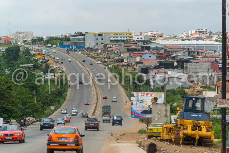 Africa photo — Vue aérienne d’une large route urbaine avec plusieurs voitures en circulation, des bâtiments colorés de chaque côté, un engin de chanti