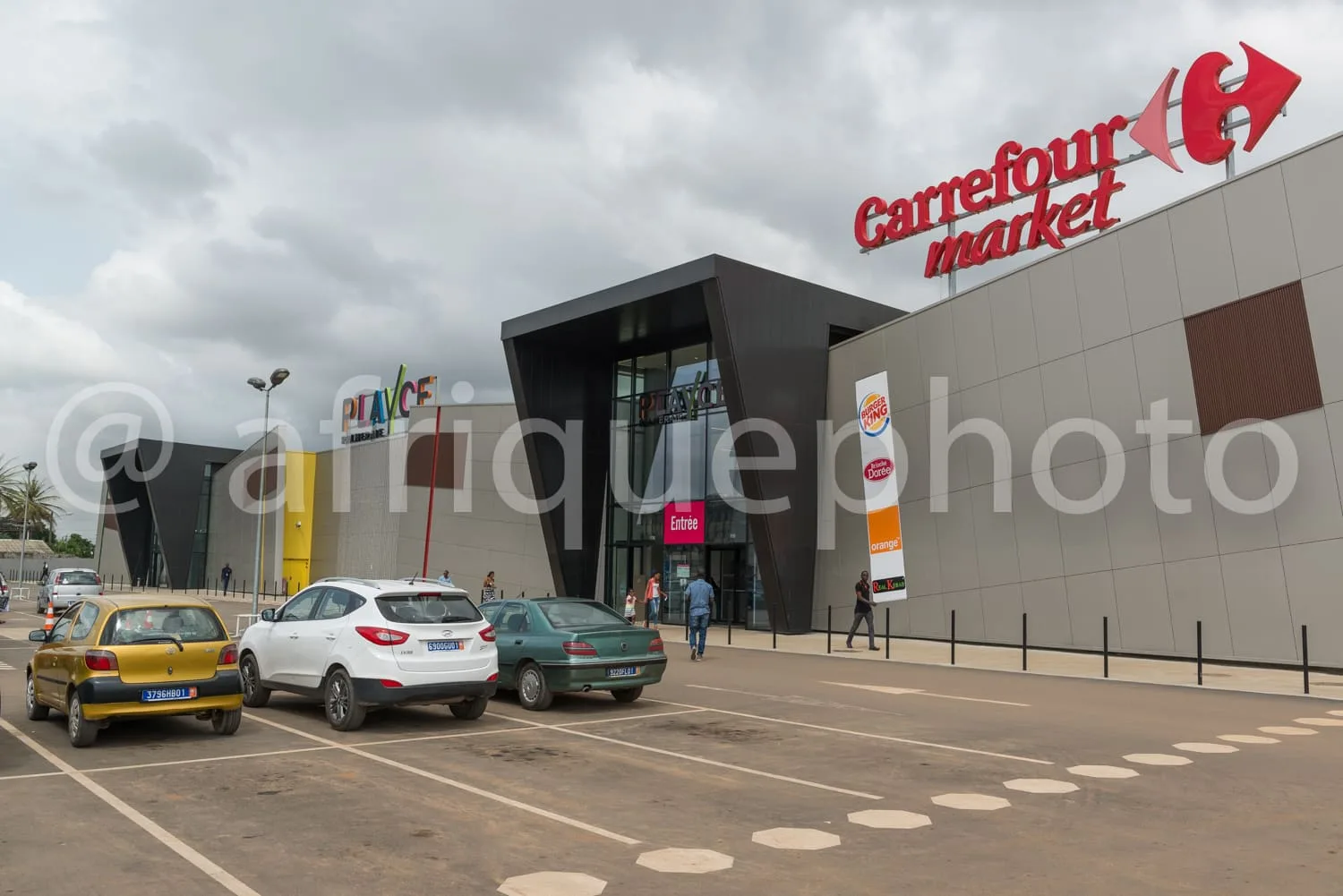 Africa photo — Image montrant l’entrée d’un supermarché Carrefour Market avec un parking devant où sont garées trois voitures de couleurs différentes.