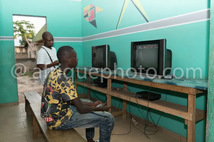Africa photo — Deux personnes assises sur un banc en bois devant deux téléviseurs anciens, jouant à des jeux vidéo. L’un porte un t-shirt à motifs col
