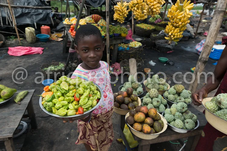 Africa photo — Un enfant souriant, vêtu d’un t-shirt à motifs, tient un plateau rempli de fruits exotiques verts et bruns, comme des avocats et des fr