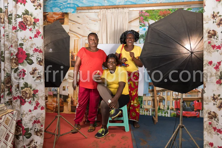 Africa photo — Trois femmes souriantes posant ensemble dans un studio photo décoré de motifs floraux, éclairées par des parapluies réflecteurs et un f