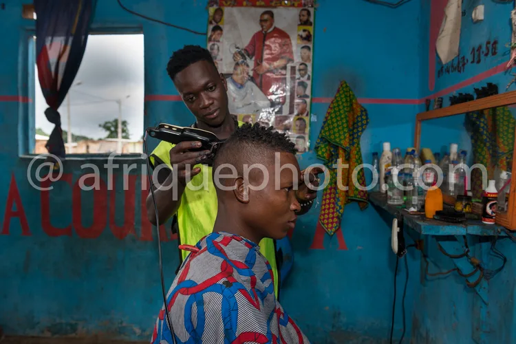 Africa photo — Un homme assis reçoit une coupe de cheveux courte d'un coiffeur dans un salon aux murs bleus, décoré de photos familiales et d'objets a