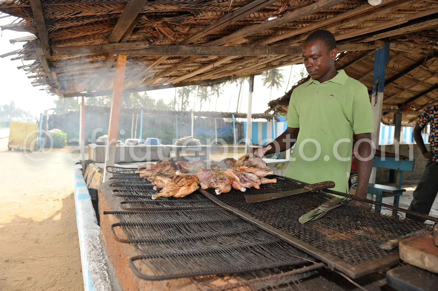 Africa photo — Un homme en polo vert prépare des morceaux de viande grillés sur un grand barbecue en métal, sous une structure en bois ouverte, avec d