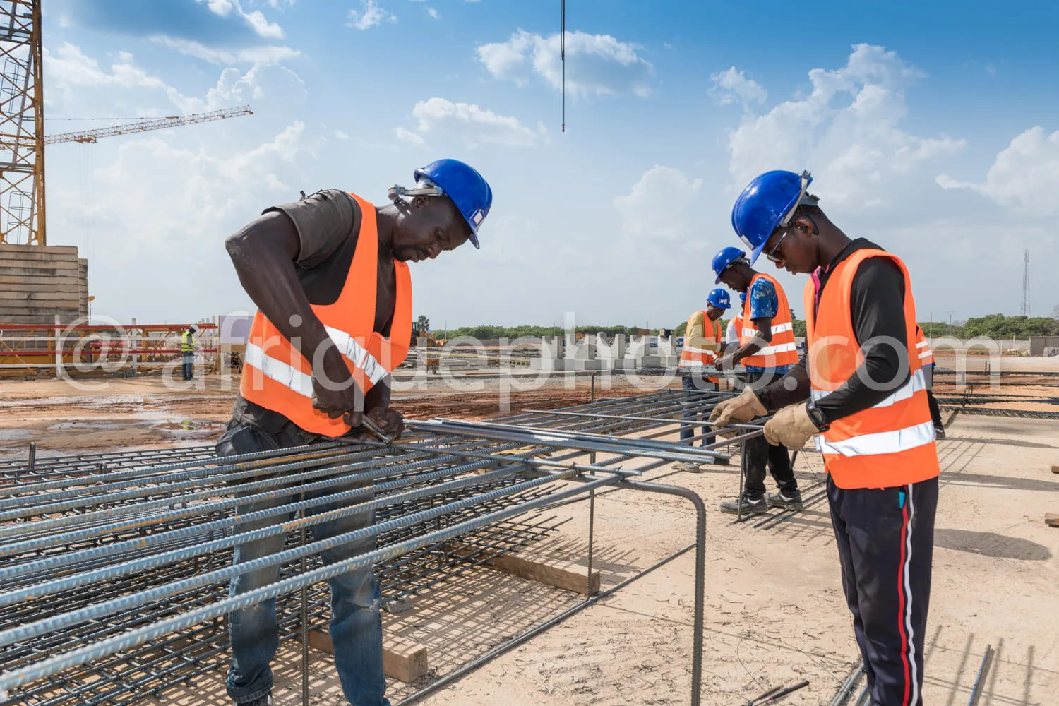 Africa photo — Quatre ouvriers équipés de casques bleus et gilets orange réfléchissants travaillent ensemble sur un chantier. Ils manipulent des armat