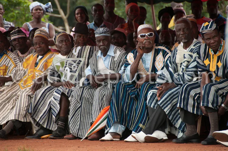 Africa photo — Un groupe d’hommes âgés assis côte à côte, vêtus de robes traditionnelles à motifs rayures et couleurs vives, portant des coiffures et