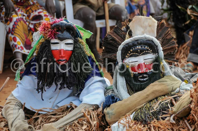 Africa photo — Deux masques traditionnels africains aux expressions marquées, assis sur des sièges en paille, entourés de tissus colorés et de motifs