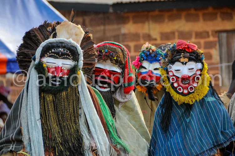 Africa photo — Quatre personnes portant des masques traditionnels aux expressions marquées, décorés de plumes, perles et tissus colorés, dans un cadre
