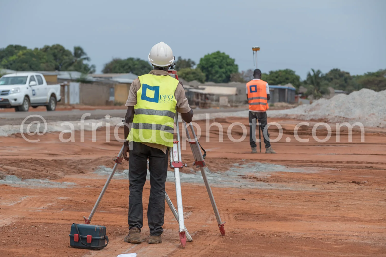 Africa photo — Un professionnel en gilet jaune et casque blanc utilise un niveau optique pour effectuer des relevés sur un sol en terre battue, avec u
