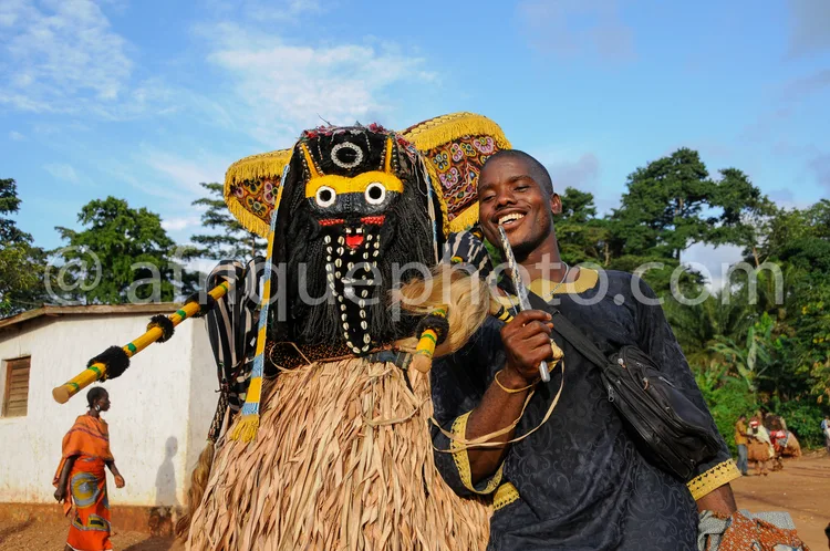 Africa photo — Un homme souriant, vêtu d'une tenue noire, pose à côté d'un masque traditionnel africain imposant aux yeux exorbités, décoré de motifs
