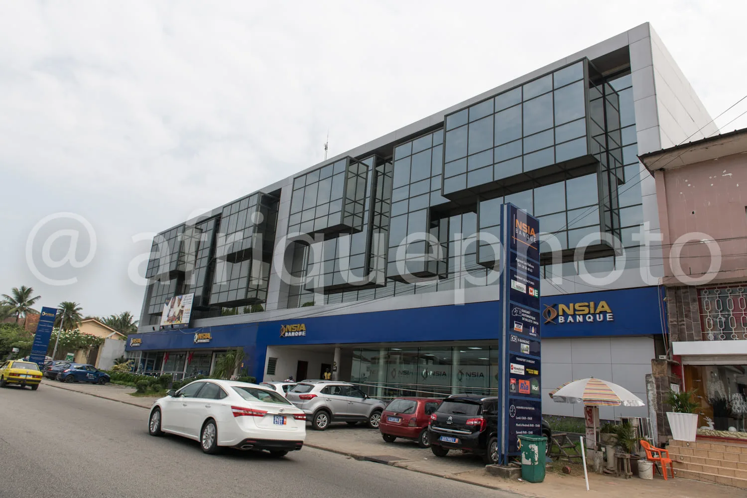 Africa photo — Un bâtiment moderne en façade vitrée avec le logo NSIA Banque en bleu et blanc, situé en bordure de route. Plusieurs voitures sont garé