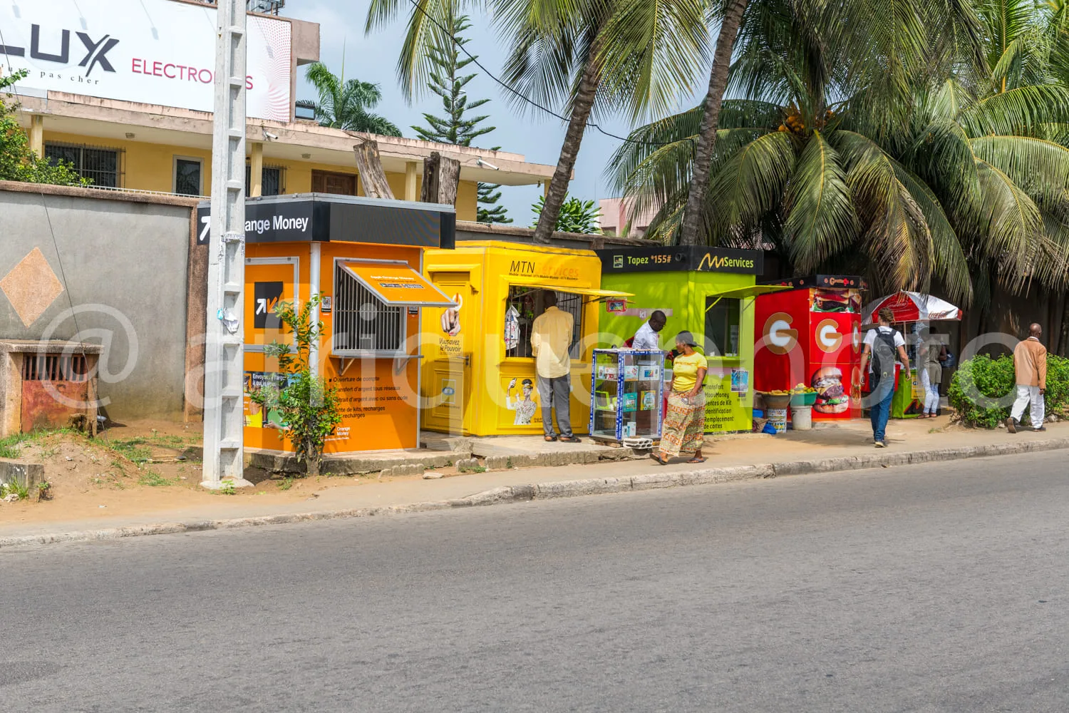Africa photo — Une rue ensoleillée avec des kiosques colorés proposant des services financiers comme Orange Money et MTN. Deux personnes discutent prè