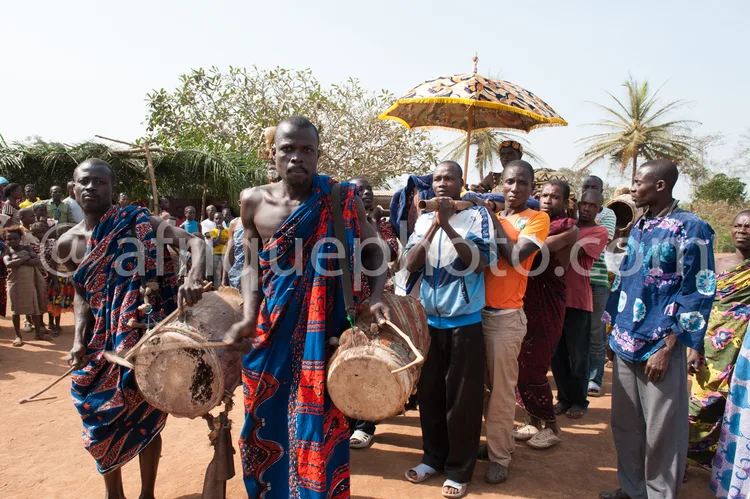 Africa photo — Un groupe de personnes défilant en plein air, vêtues de tenues traditionnelles colorées et ornées de motifs, portant des tambours en pe