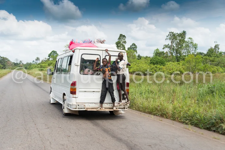 Africa photo — Un minibus blanc circulant sur une route asphaltée avec quatre passagers debout à l’arrière, dont un enfant, sous un ciel bleu et des a