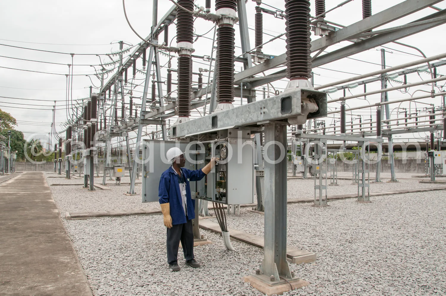 Africa photo — Un technicien en uniforme bleu inspecte et manipule un boîtier électrique fixé à une structure métallique dans un poste haute tension,