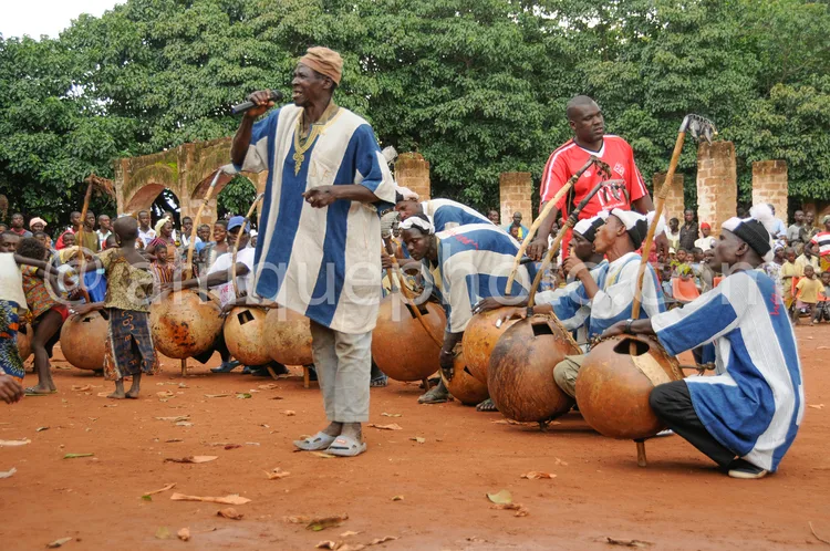 Africa photo — Un groupe de musiciens en tenue traditionnelle, dont certains portent des vêtements bleus et blancs, jouent des tambours en terre cuite