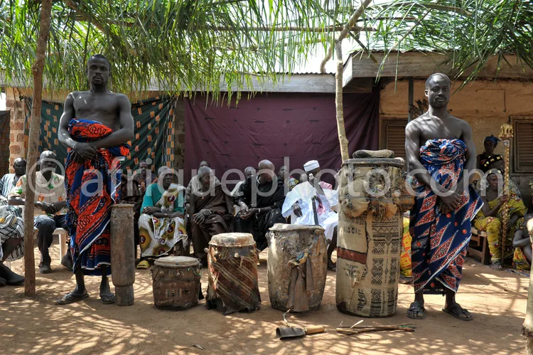 Africa photo — Deux hommes vêtus de pagnes colorés et ornés de motifs traditionnels se tiennent devant des tambours en bois sculpté et des objets ritu