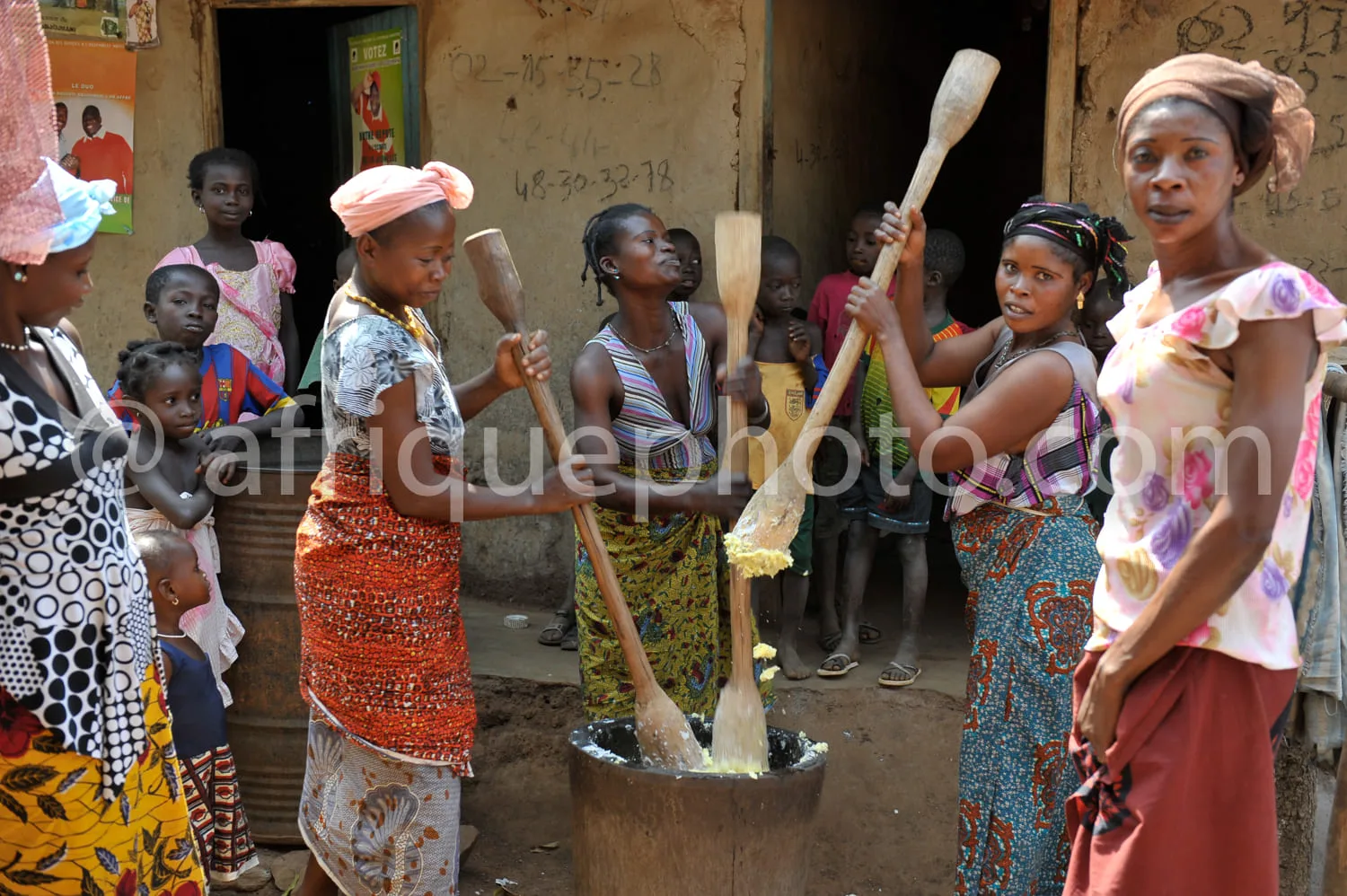 Africa photo — Un groupe de femmes en tenues colorées et coiffures variées se tient autour d’un grand récipient en métal. Elles mélangent et pétrissen
