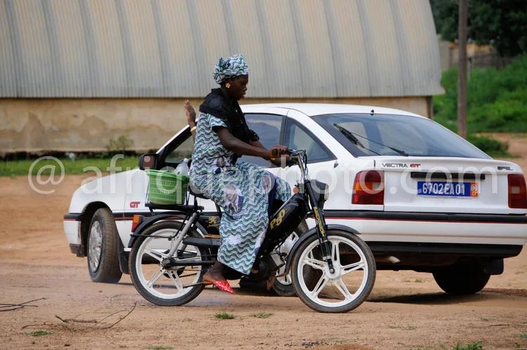Africa photo — Une femme vêtue d’une robe colorée à motifs et d’un foulard sur la tête conduit une moto légère, avec une Opel Vectra blanche stationné