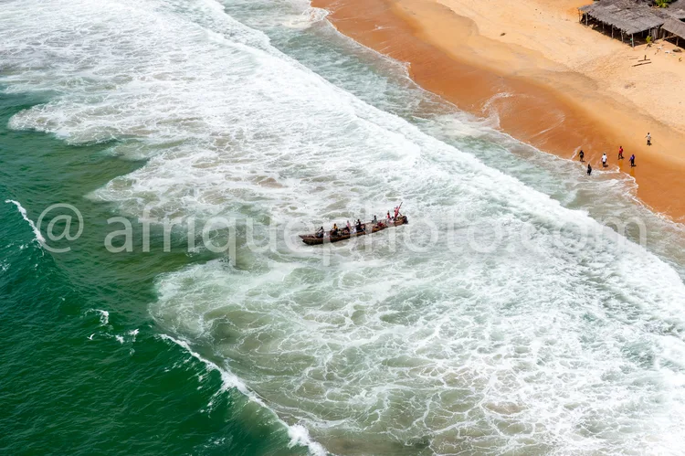 Africa photo — Image montrant une pirogue avec des pêcheurs naviguant près du rivage, avec des vagues puissantes en mer et une plage de sable clair où