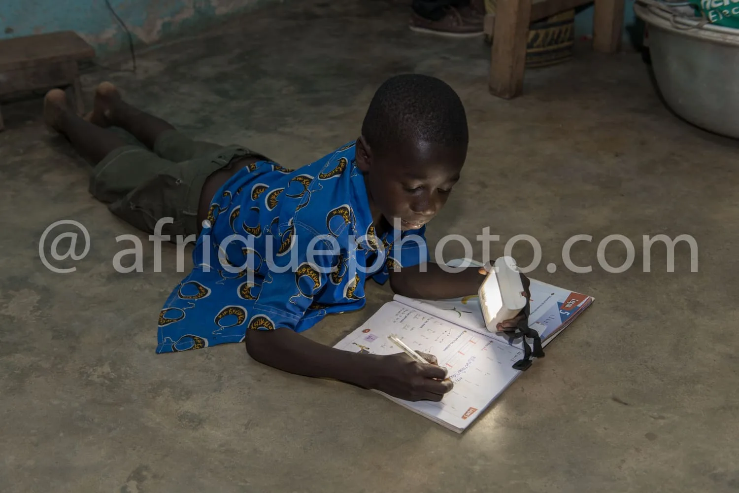 Africa photo — Un enfant assis sur le sol, concentré sur un cahier éclairé par une lampe torche, dans un environnement modeste avec des objets du quot