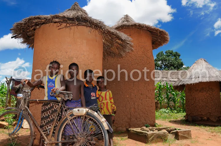 Africa photo — Trois enfants souriants posant devant une case en terre crue aux toits de chaume, avec un vélo à côté, dans un village traditionnel sou