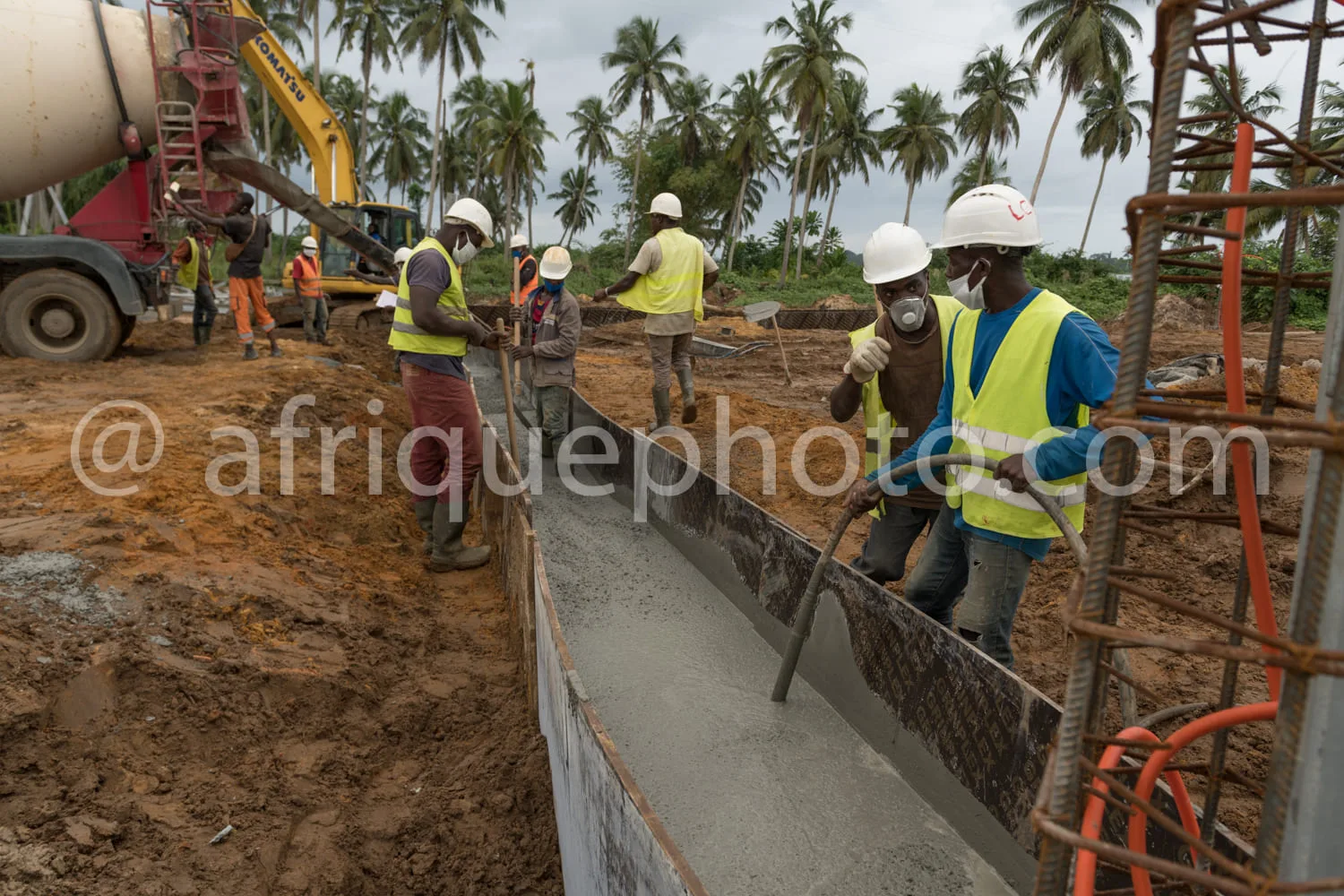 Africa photo — Six ouvriers en tenue de sécurité, casques et gilets, versent manuellement du béton frais dans une longue tranchée préparée, entourés d
