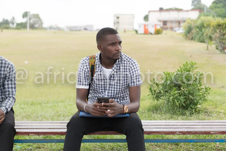 Africa photo — Un homme assis sur un banc en bois dans un parc vert, portant un t-shirt à carreaux et un pantalon noir, regardant attentivement son sm
