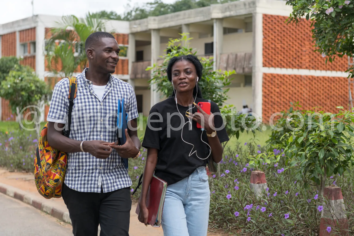 Africa photo — Deux personnes souriantes marchent côte à côte sur un chemin bordé de fleurs et de bâtiments universitaires. L’une porte un sac coloré
